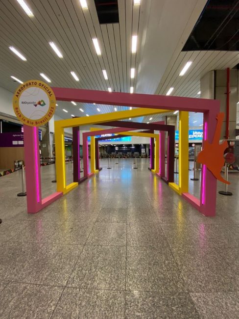 Portal cenográfico para o Rock in Rio no Aeroporto Rio Galeão, com molduras coloridas em rosa e amarelo no saguão