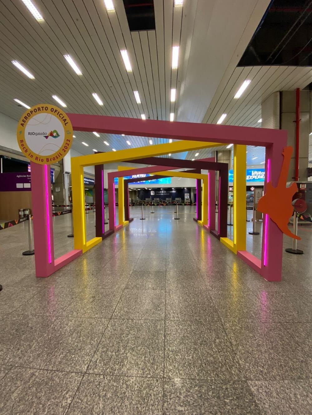 Portal cenográfico para o Rock in Rio no Aeroporto Rio Galeão, com molduras coloridas em rosa e amarelo no saguão