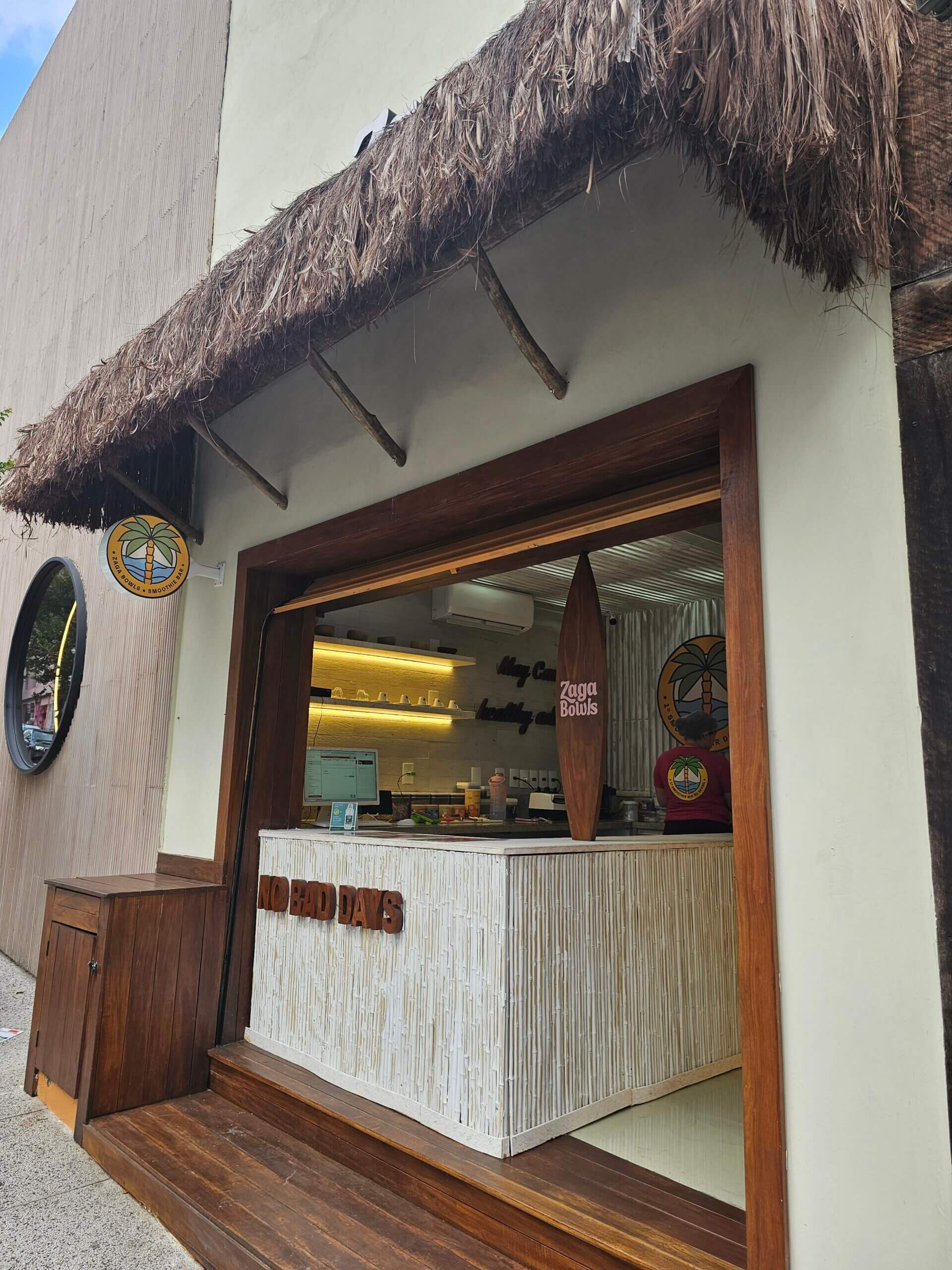 Fachada do bar Zaga Bowls em São Paulo, com cobertura de palha, balcão de madeira clara e acabamento rústico