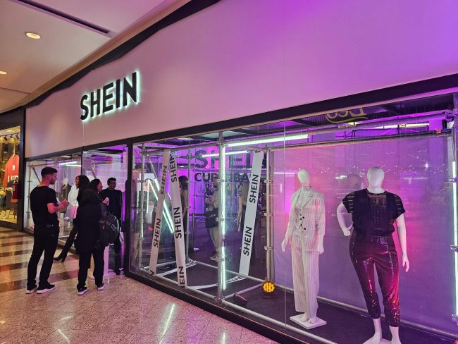 Fachada da loja pop-up Shein em shopping, com vitrine de vidro e manequins iluminados