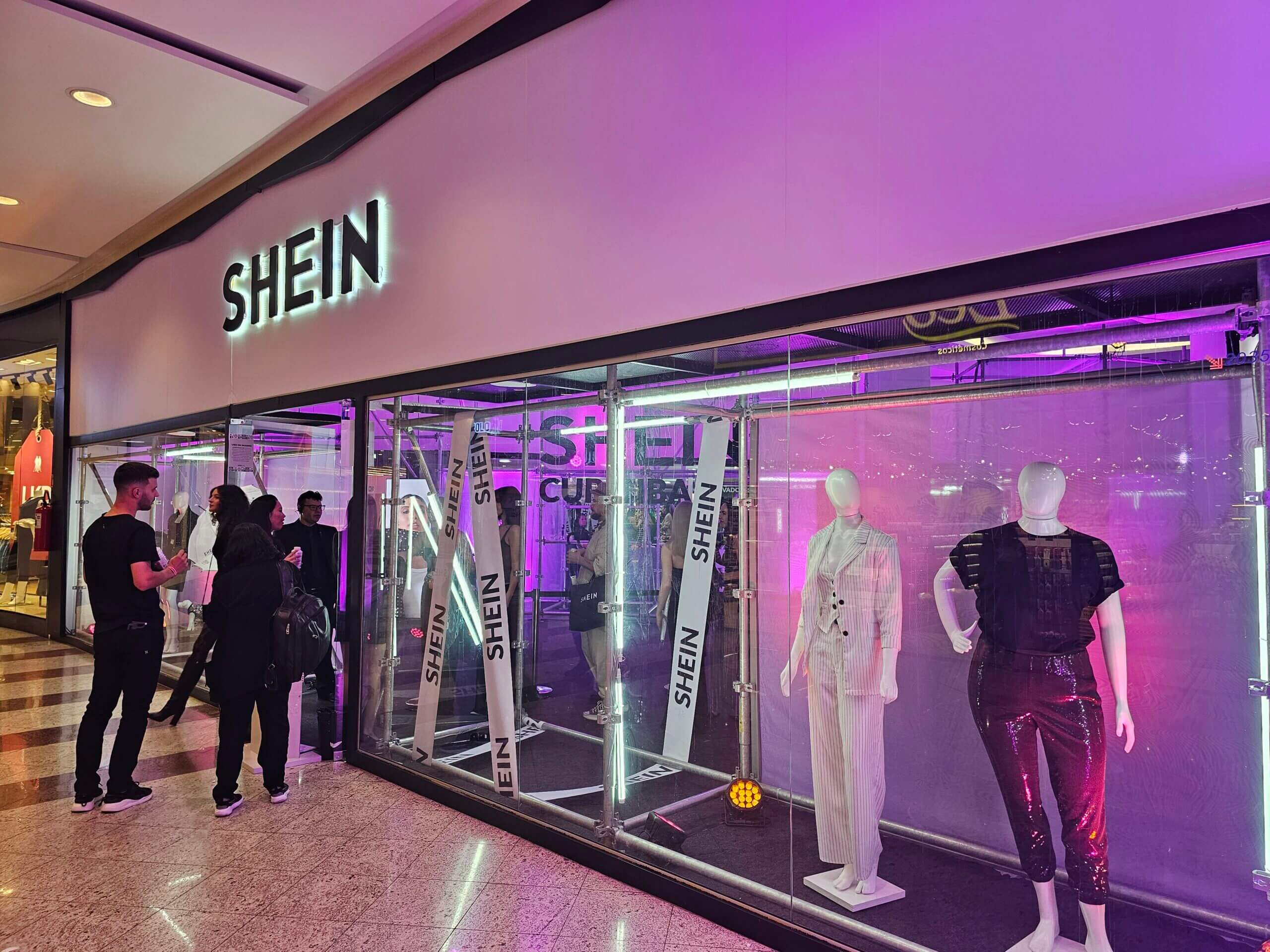 Fachada da loja pop-up Shein em shopping, com vitrine de vidro e manequins iluminados