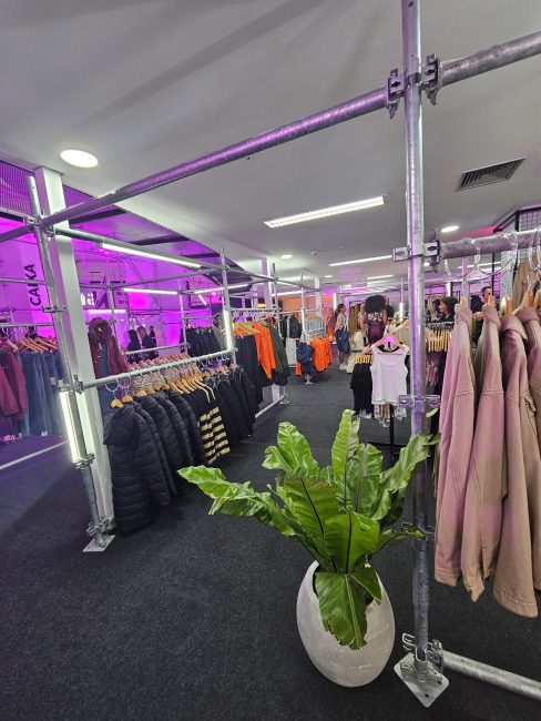 Área de vendas da pop-up Shein com araras de roupas, planta e iluminação roxa