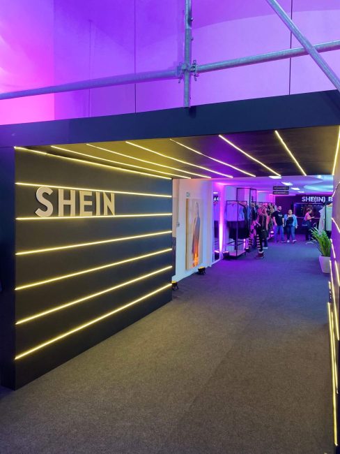 Corredor escuro com luminárias lineares douradas e logo Shein