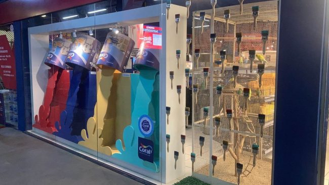 Vitrine Coral na loja Vilarejo em Cabo Frio, com latas de tinta derramando cores vermelha, azul, amarela e verde