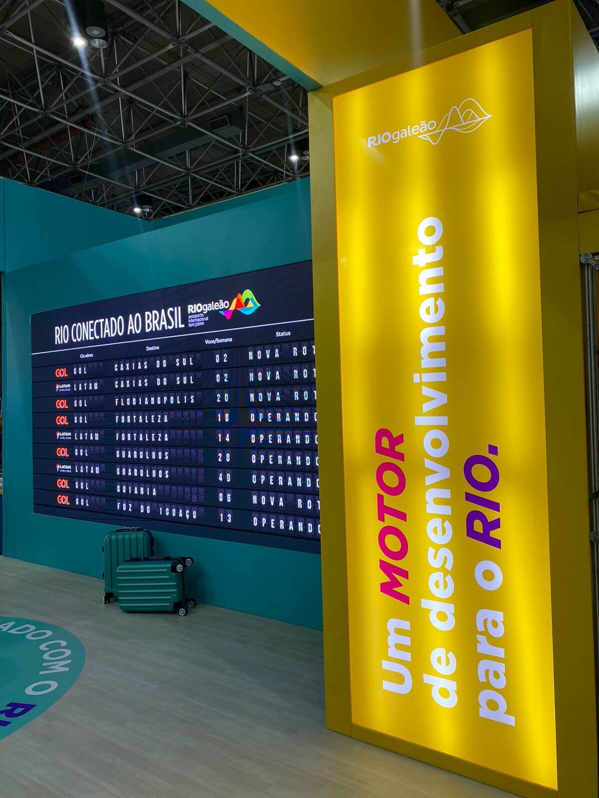 Painel luminoso amarelo com a frase "Um motor de desenvolvimento para o Rio" e painel de voos no stand Rio Galeão na Feira ABAV 2023