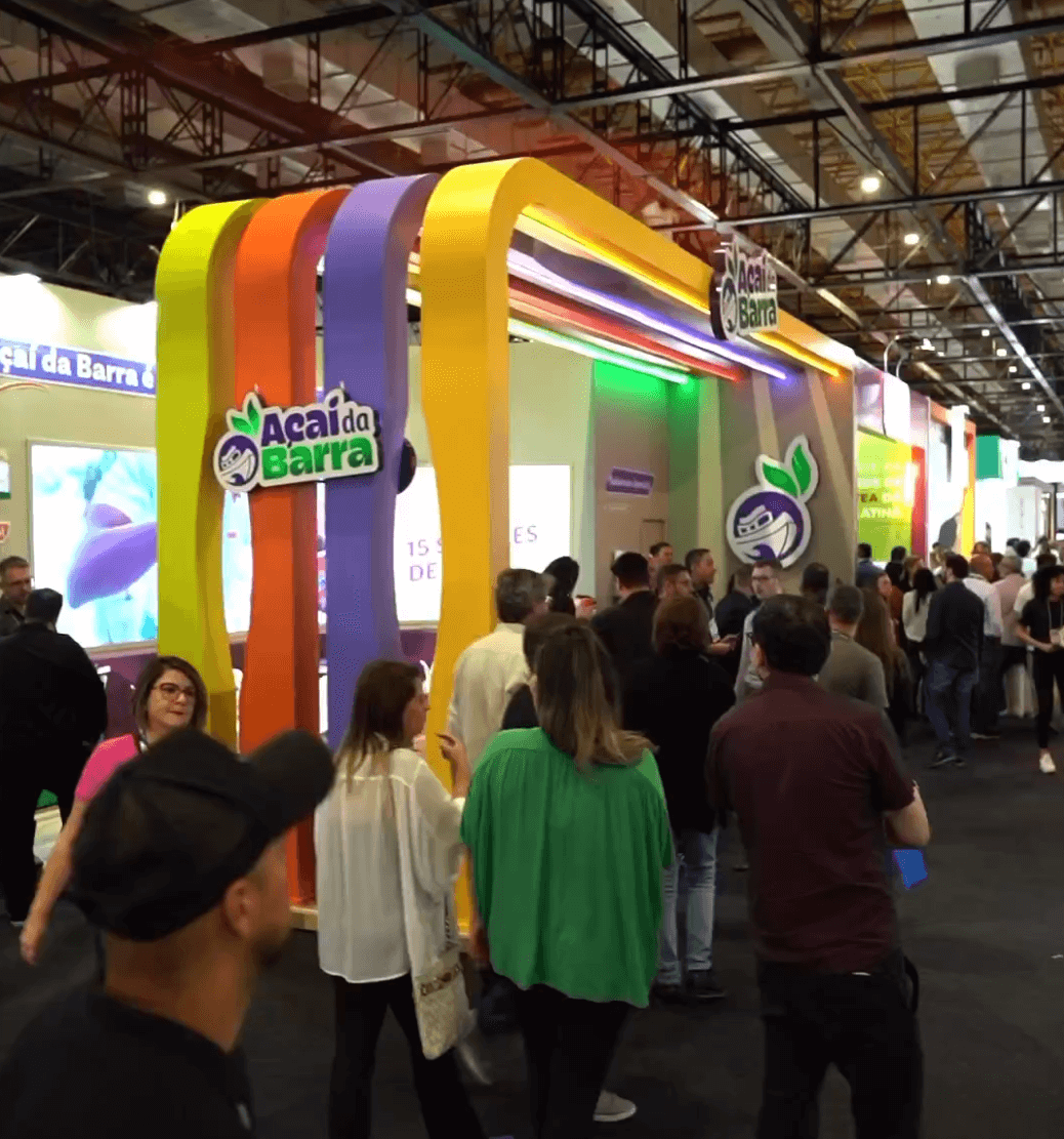 Stand do Açaí da Barra na feira da ABF 2023, com arco de faixas coloridas em amarelo, laranja e roxo e visitantes passando