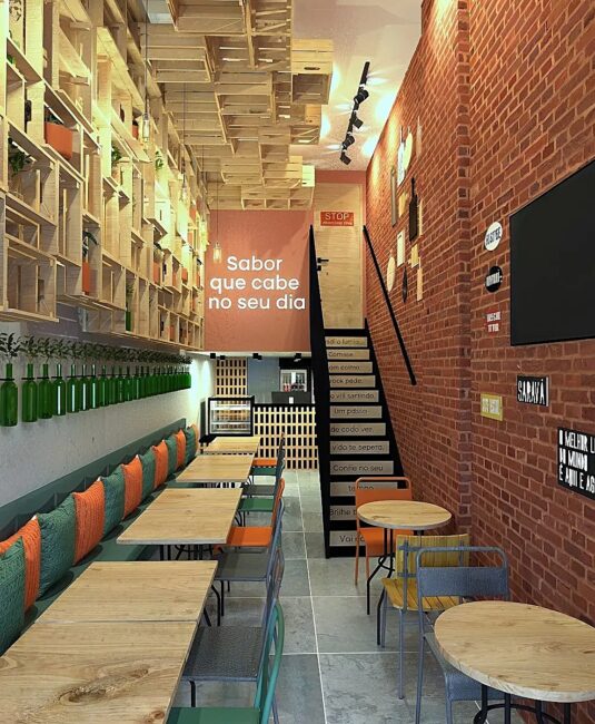 Interior do restaurante Vanilla Café & Comida com estantes de caixotes, garrafas verdes e banco com almofadas, projeto da Unpack Design